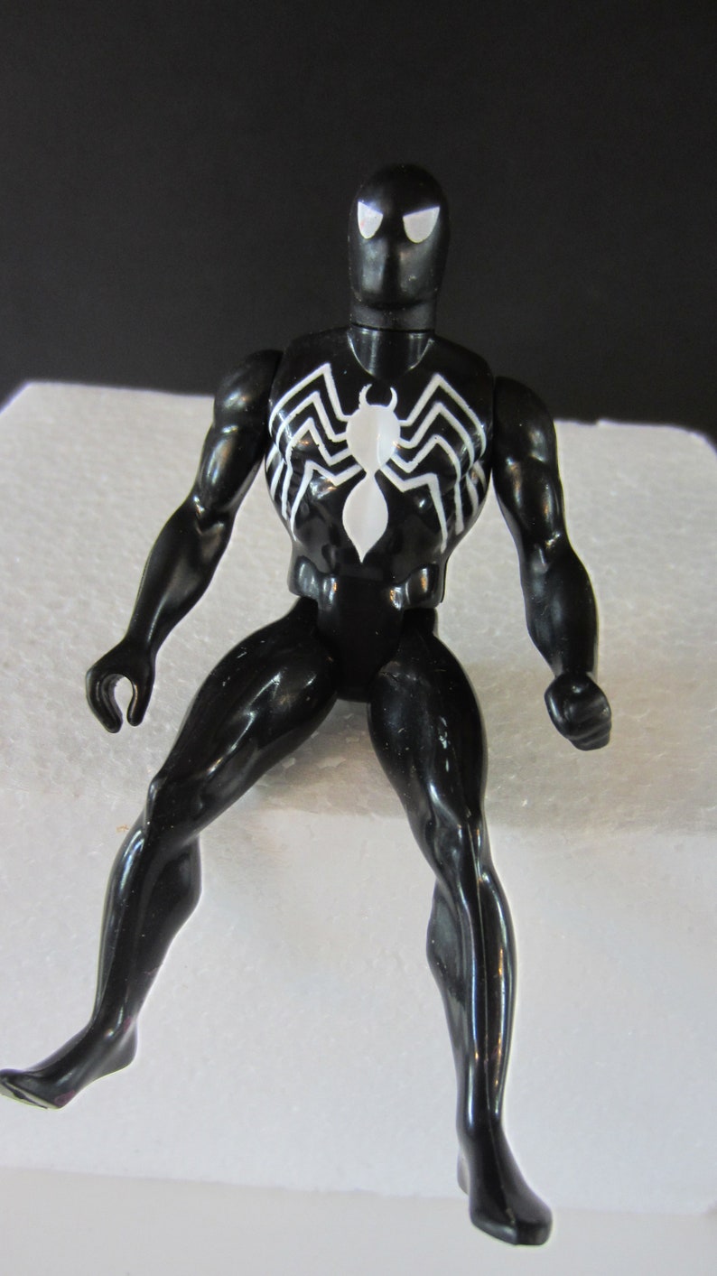 May include: A black action figure of a superhero with a white spider emblem on the chest. The figure has white eye shapes and is posed with one leg extended. The figure is glossy and appears to be made of plastic.