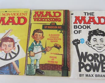 Może przedstawiać: Trzy książki z magazynu MAD w stylu vintage. Okładki przedstawiają kultową postać Alfreda E. Neumana w różnych ilustracjach. Tytuły to "The Non-Violent MAD", "MAD Vertising" i "The MAD Book of Word Power". Książki są w dobrym stanie.