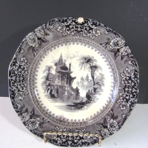 May include: Antique decorative plate with a black and white Asian-inspired landscape scene. The plate has a gray border with floral and scrollwork designs. The plate is displayed on a gold-colored metal stand.