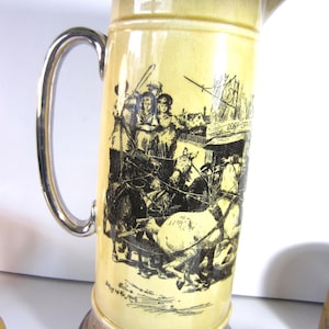 May include: A tall, yellow ceramic pitcher with a silver handle and rim. The pitcher features a black and white illustration of a horse-drawn carriage and people. The base of the pitcher is silver with a decorative pattern.
