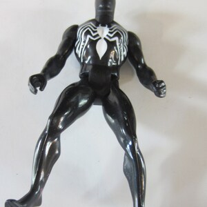 May include: A black action figure of a superhero with a white spider emblem on the chest. The figure is posed with legs spread and arms out, showcasing a glossy finish. The figure is approximately 6 inches tall.