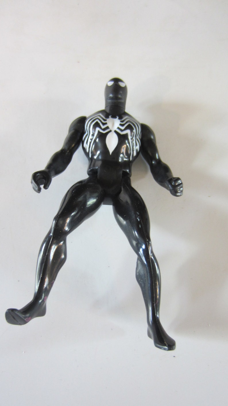 May include: A black action figure of a superhero with a white spider emblem on the chest. The figure is posed with legs spread and arms out, showcasing a glossy finish. The figure is approximately 6 inches tall.