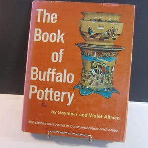 May include: An orange book titled "The Book of Buffalo Pottery" with white text. The cover features a colorful illustration of two pottery pieces. The book is by Seymour and Violet Altman, with 400 pieces illustrated.