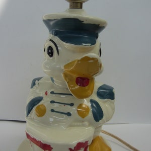 May include: A vintage ceramic lamp featuring a duck character in a sailor's hat and uniform. The lamp has a white base with red and yellow accents, and a brass lamp holder. The duck is holding a drum.