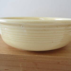 May include: A pale yellow ceramic bowl with a rounded shape and a slightly flared rim. The bowl has horizontal ridged detailing around the exterior. The bowl is sitting on a wooden surface. This bowl is suitable for serving food.