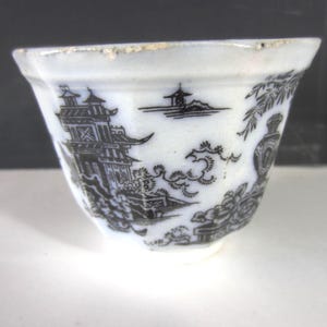 May include: A white teacup with a black and white Asian-inspired design. The cup features a detailed landscape scene with pagodas, trees, and clouds. The rim of the cup has a slightly irregular shape.