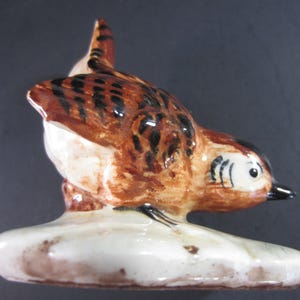 May include: A ceramic bird figurine with brown and black markings, perched on a white base. The bird has a detailed design, with a black beak and eye details. The figurine is likely a decorative item.