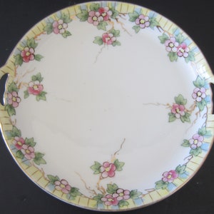 May include: A round, off-white ceramic serving plate with two handles. The rim is decorated with a floral pattern of pink flowers, green leaves, and a yellow and blue border. The plate has a gold trim.