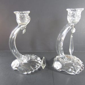 May include: A pair of clear glass candle holders. Each holder features a curved stem, a seashell-shaped base, and a decorative crystal pendant. The candle cups have a floral design. The holders are approximately 8 inches tall.