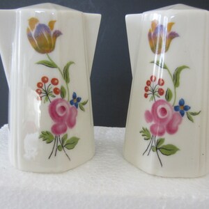 May include: A pair of vintage salt and pepper shakers. Each shaker is white with a floral design featuring pink roses, orange berries, blue flowers, and a yellow and purple tulip. The shakers have a unique, angular shape.