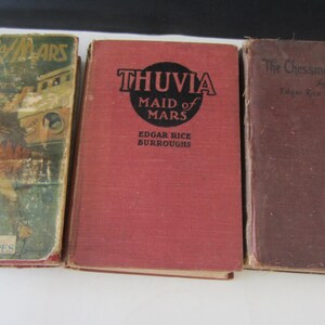 May include: Three vintage books with worn covers. The titles include "The Gods of Mars", "Thuvia, Maid of Mars", and "The Chessmen of Mars" by Edgar Rice Burroughs. The books have a retro aesthetic.