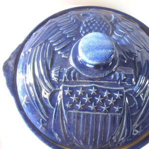 May include: A blue ceramic casserole dish lid with an embossed design featuring an eagle, shield, and stars. The lid has a round knob and handles. The design is a patriotic theme.