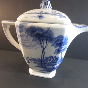 May include: A white and blue ceramic teapot with a lid. The teapot features a landscape scene with trees, a house, and a boat. The handle and spout are white. The lid has a decorative finial.