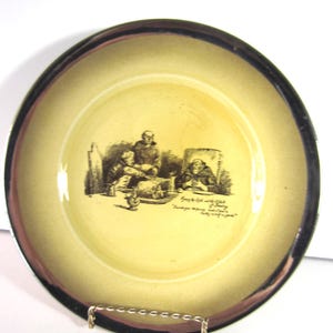 May include: A vintage ceramic plate with a yellow center and a dark brown rim. The plate features a black and white illustration of people in a room, with text below the image. The plate is displayed on a silver wire stand.