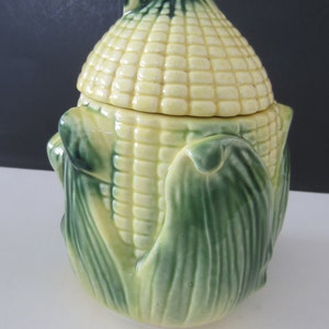 May include: A ceramic corn-shaped container with a lid. The container is primarily yellow with a textured surface, mimicking corn kernels. Green leaves form the base and the top of the container, with a dark green accent on the lid's handle. The container is designed to resemble an ear of corn.