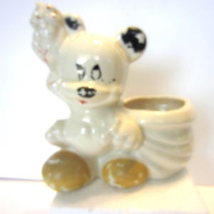 May include: A vintage ceramic planter in the shape of a cartoon bear. The bear is primarily off-white with black accents on the ears and eyes, and yellow feet. The planter has a small opening for plants or other decorative items.