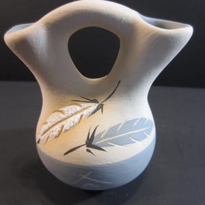May include: A cream-colored ceramic vase with a round opening and flared top. The vase features a light blue base and feather designs in cream and blue, with black accent lines. The vase is approximately 6 inches tall.