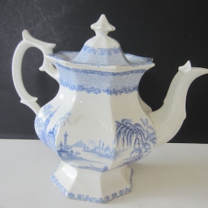 May include: A white and blue ceramic teapot with a lid, spout, and handle. The teapot features a blue landscape design and decorative blue trim. The teapot is likely vintage and is on a white surface.