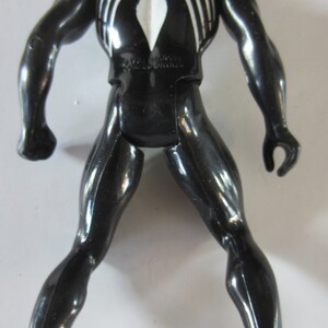 May include: Black action figure of a superhero with white spider-web-like markings on the chest and back. The figure has a muscular build and is posed with legs apart and arms slightly outstretched. The figure is made of a glossy plastic material.