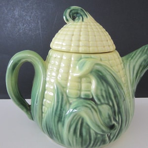 May include: A ceramic teapot shaped like an ear of corn, with a light yellow body and a green base and handle. The lid is also yellow and shaped like the top of the corn. The teapot has a spout and a handle, and is designed to look like a complete ear of corn.