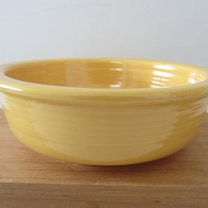 May include: A yellow ceramic bowl with a rounded rim. The bowl is a solid, pale yellow color and sits on a wooden surface. The bowl is empty and appears to be new.