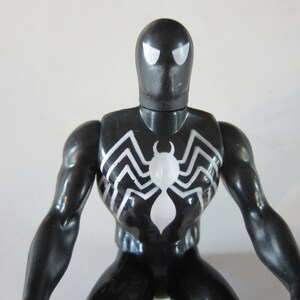 May include: A black Spider-Man action figure with white spider emblem on the chest. The figure has white eyes and a glossy finish. The figure is a collectible toy.