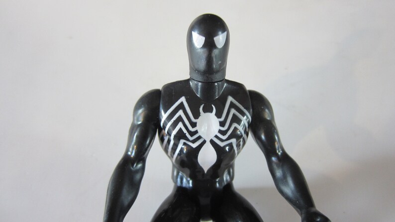 May include: A black Spider-Man action figure with white spider emblem on the chest. The figure has white eyes and a glossy finish. The figure is a collectible toy.