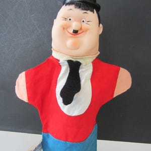 May include: A vintage hand puppet of a cartoon character, featuring a round face, black bowler hat, and a red shirt with a white bib and black tie. The puppet's lower half is blue. A tag is attached to the side.