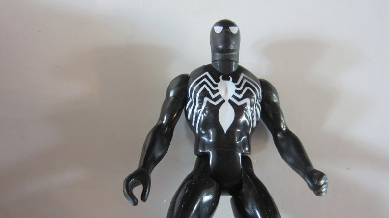 May include: A black action figure of a superhero with a white spider emblem on the chest. The figure has a black head with white eye shapes and white spider web designs on the chest and back.