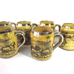 May include: Set of six vintage ceramic mugs with a yellow and black landscape design. Each mug has a silver-colored handle and rim. The mugs feature a horse-drawn carriage scene.