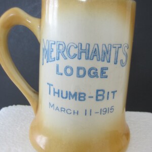 May include: A vintage ceramic mug with a handle, featuring the text "MERCHANTS LODGE THUMB-BIT MARCH 11-1915" in blue lettering. The mug has a cream-colored base with a gradient of tan around the top and bottom.