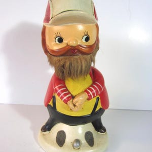 May include: A vintage ceramic figurine of a bearded man wearing a cap and red jacket with white stripes. The figurine has a yellow shirt, black pants, and a cream-colored base with dark spots.