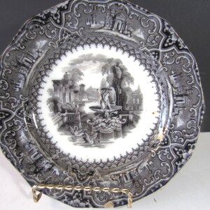 May include: A black and white decorative plate with a scalloped edge. The plate features a detailed landscape scene with a fountain, trees, and buildings. The border has an ornate, repeating pattern. The plate is displayed on a gold-colored wire stand.