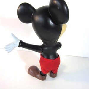 Pode incluir: Uma figura vintage do Mickey Mouse, vista por tr&aacute;s, com cabe&ccedil;a e orelhas pretas, rosto amarelo e cal&ccedil;&otilde;es vermelhos. A figura tem luvas brancas e sapatos castanhos. A figura est&aacute; em pose com um bra&ccedil;o estendido.