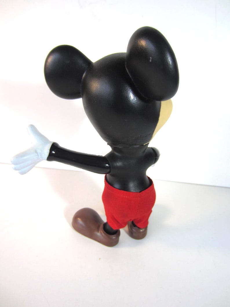Pode incluir: Uma figura vintage do Mickey Mouse, vista por tr&aacute;s, com cabe&ccedil;a e orelhas pretas, rosto amarelo e cal&ccedil;&otilde;es vermelhos. A figura tem luvas brancas e sapatos castanhos. A figura est&aacute; em pose com um bra&ccedil;o estendido.