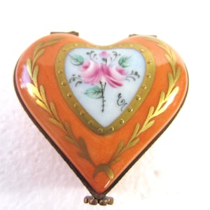 May include: Heart-shaped trinket box in orange with a floral design. The box features a central heart-shaped panel with a pink rose illustration, framed by gold trim and laurel leaves. The box has a small, decorative clasp.