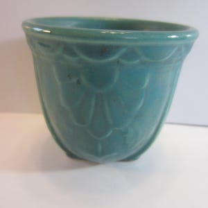 May include: A teal ceramic planter with a scalloped design. The small, rounded planter has a slightly flared rim and sits on three small feet. The teal color is consistent throughout the planter.