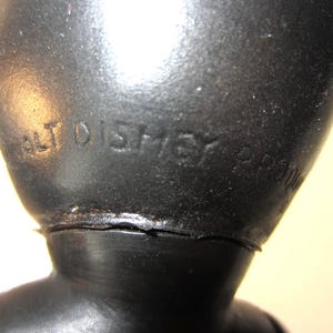 Pode incluir: Close-up de uma figura vintage preta com o texto em relevo "WALT DISNEY" vis&iacute;vel. A figura parece ser feita de um material duro, possivelmente pl&aacute;stico. A superf&iacute;cie tem uma apar&ecirc;ncia ligeiramente texturizada.