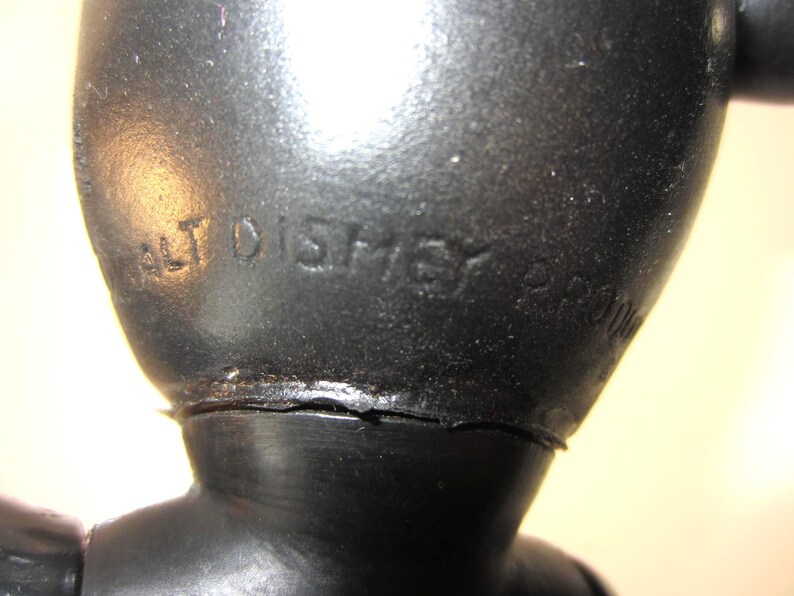 Pode incluir: Close-up de uma figura vintage preta com o texto em relevo "WALT DISNEY" vis&iacute;vel. A figura parece ser feita de um material duro, possivelmente pl&aacute;stico. A superf&iacute;cie tem uma apar&ecirc;ncia ligeiramente texturizada.