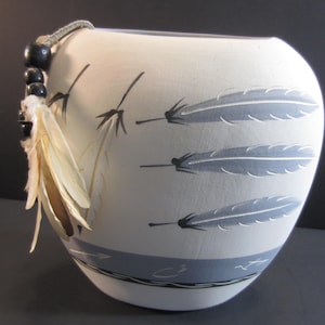 May include: A white ceramic bowl with a gray and black feather design. The bowl has a light blue band around the middle and is adorned with a beaded string and feathers.