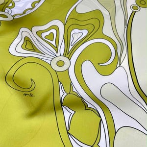 May include: A smooth fabric scarf featuring a bold, abstract floral pattern in shades of lime green, white, and black. The design consists of large, flowing organic shapes, curved lines, and circular motifs with a signature printed on the fabric.