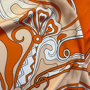 Op de afbeelding: Een gladde stof met een opvallend grafisch patroon in levendig oranje, zacht crème en wit. Het ontwerp bestaat uit vloeiende, gebogen lijnen, geometrische vormen en gestileerde bloemmotieven die zich over het oppervlak herhalen.