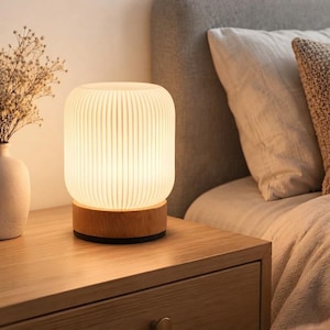 May include: A cylindrical table lamp with a white, vertically ribbed, translucent shade and a circular light-toned wooden base. The lamp emits a warm, soft glow, illuminating the surrounding area with a gentle light, creating a calm atmosphere.