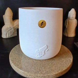 May include: A matte white cylindrical ceramic planter with a smooth finish. The front features a small, circular gold sticker with a black bird silhouette and text, and a subtle, embossed relief of a map shape near the base.