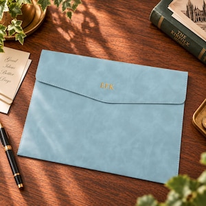 May include: A light blue document folder with a smooth, matte finish and a pointed flap closure. The centre of the flap features the gold-coloured embossed letters EFK. The folder has visible stitching along the edges and a rectangular shape.