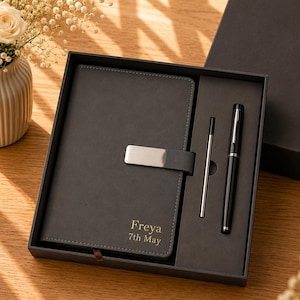 May include: A dark gray notebook with a silver metal clasp and the gold text Freya 7th May, presented in a black gift box alongside a black ballpoint pen and a silver metal ink refill. The notebook features a smooth cover and stitched edges.