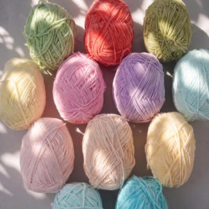 May include: A collection of eleven yarn balls in various pastel and vibrant colors including light green, coral, olive, pale yellow, pink, lavender, light blue, and cream. Each ball features a soft, textured, velvet-like chenille yarn strand.