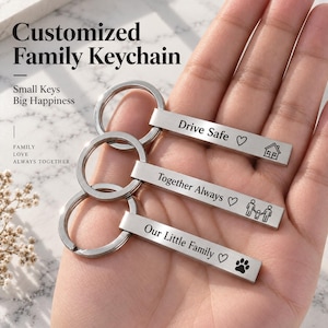 Könnte beinhalten: Drei rechteckige Schlüsselanhänger aus silberfarbenem Metall mit Schlüsselringen. Jeder Stab ist mit Text und Symbolen graviert: Drive Safe mit Herz und Haus, Together Always mit Herz und Familiensymbol sowie Our Little Family mit Herz und Pfotenabdruck.