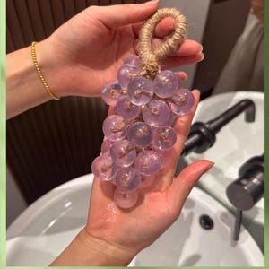 Puede incluir: Un racimo de cuentas de jabón esféricas, translúcidas y de color morado claro, dispuestas en forma de uvas. Las cuentas están unidas por un lazo de cuerda de yute gruesa de color natural en la parte superior, diseñado para colgar o sujetar.