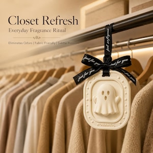May include: A cream-colored, oval-shaped hanging air freshener featuring a raised ghost figure and the word ROMANTIC at the bottom. It is suspended by a black ribbon printed with the text just for you, hanging from a closet rod among clothing.